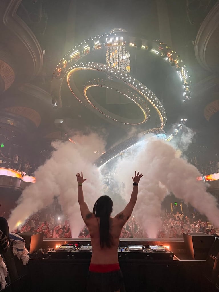 DJ at concert with arms raised above turntables, surrounded by white stage smoke and spiral LED lighting overhead